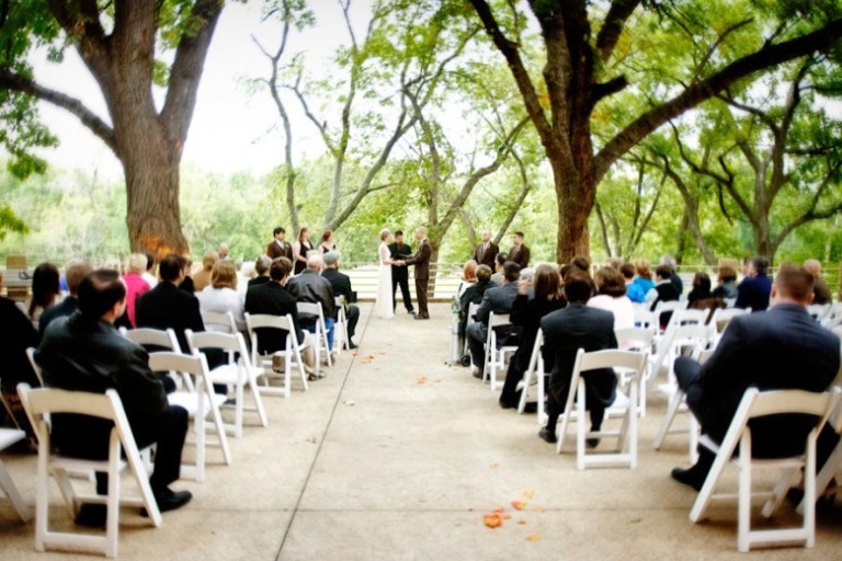 An outdoor wedding ceremony taking place under beautiful mature trees with guests seated in white chairs watching the ceremony.