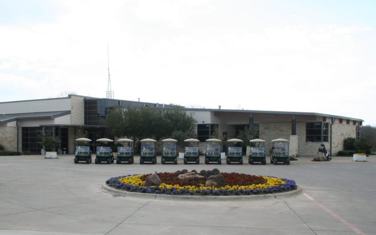 Modern clubhouse building with stone and glass architecture featuring a line of golf carts parked in front and a colorful circular flower bed in the foreground.