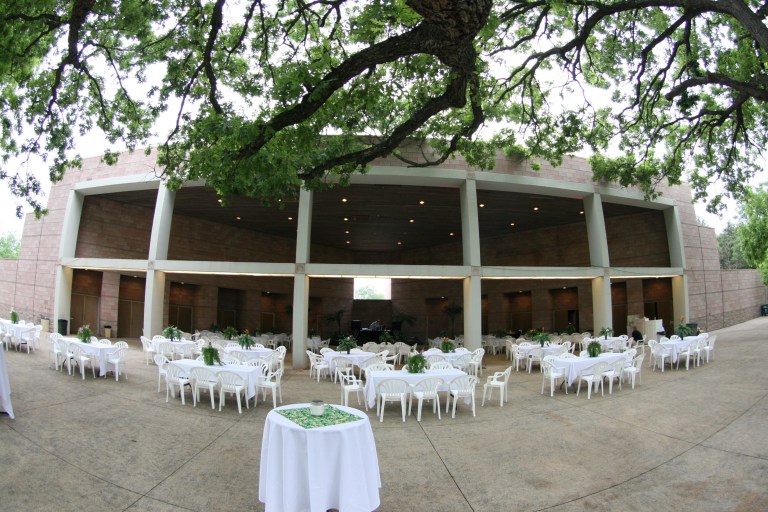 An elegant event setup under a modern pavilion with round tables, white linens, and mature trees creating a sophisticated outdoor dining atmosphere.