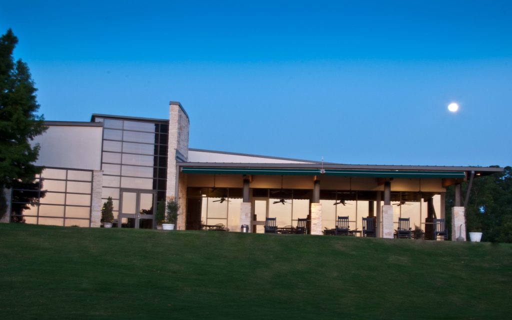 Evening twilight shot of the clubhouse with outdoor covered patio area featuring seating and ceiling fans, set against a manicured lawn with the moon visible in the blue sky.