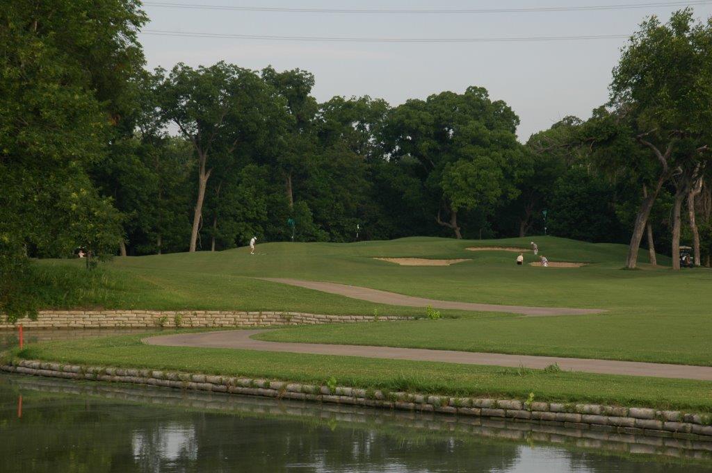 A golf green with sand bunker surrounded by mature trees with golfers visible in the distance and a water feature in the foreground.