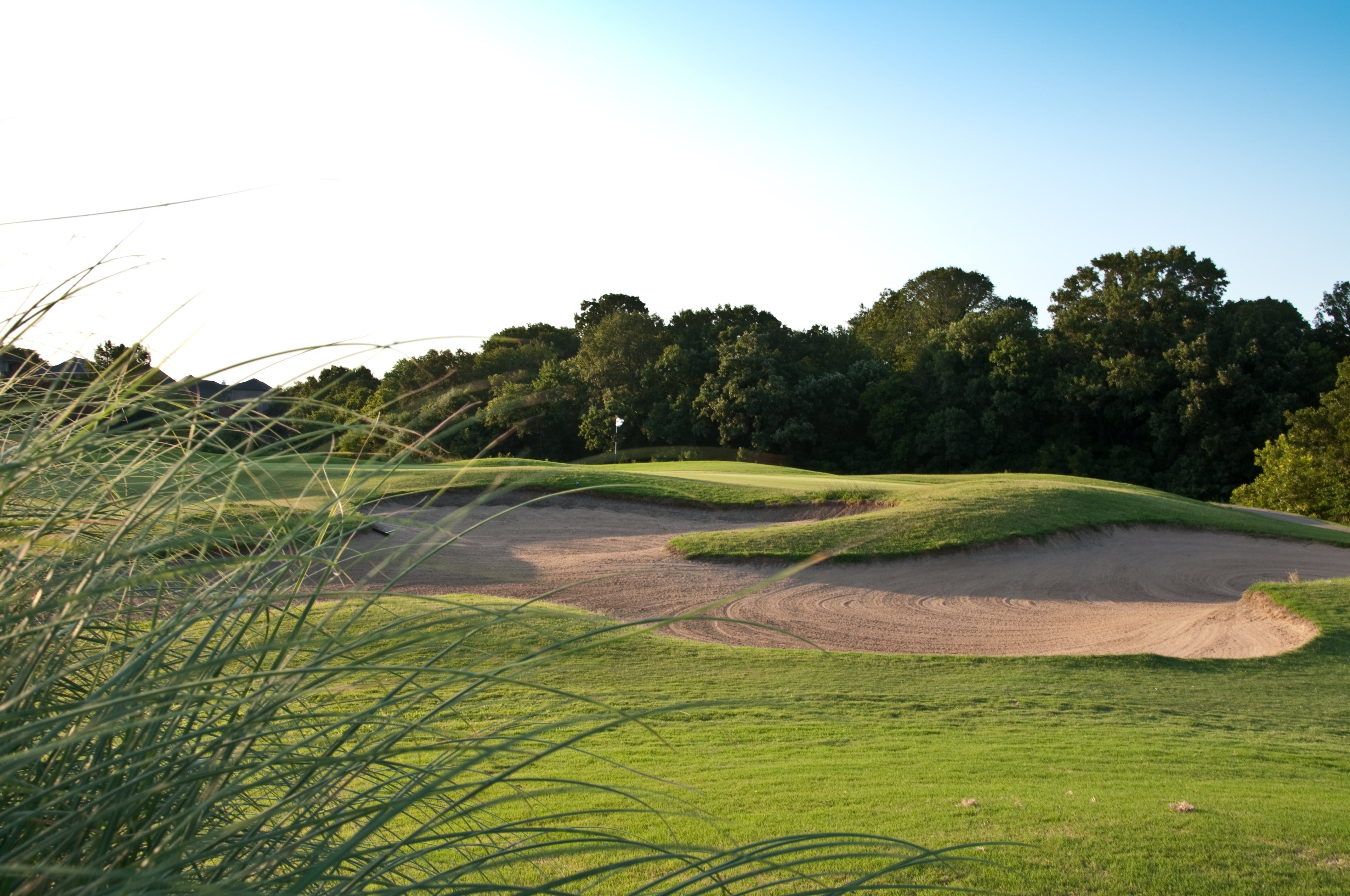 A beautifully lit golf hole featuring multiple sand bunkers and undulating green terrain surrounded by mature trees during golden hour.