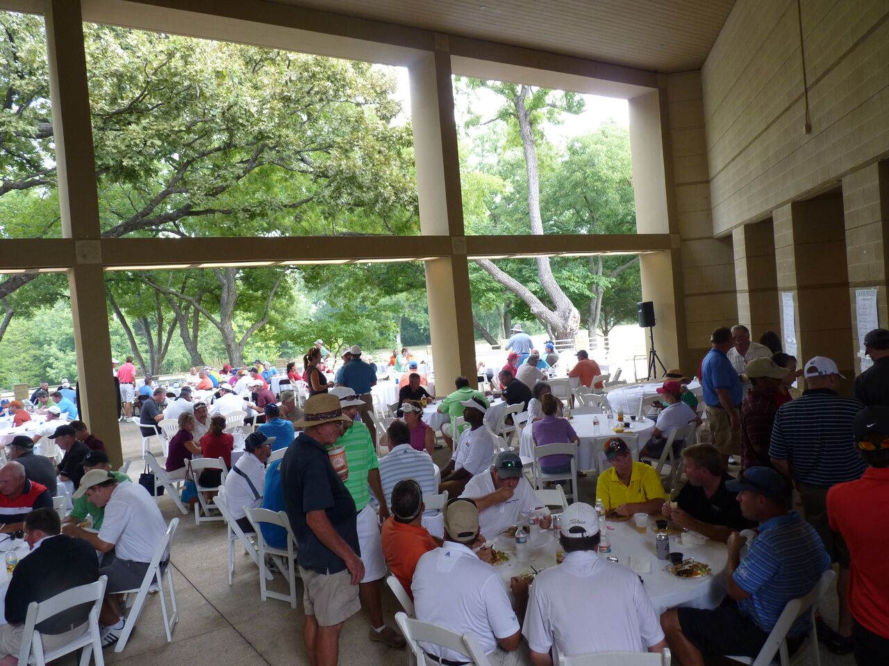 A lively golf tournament or event gathering with many people seated at tables under a covered pavilion with golf course views in the background.