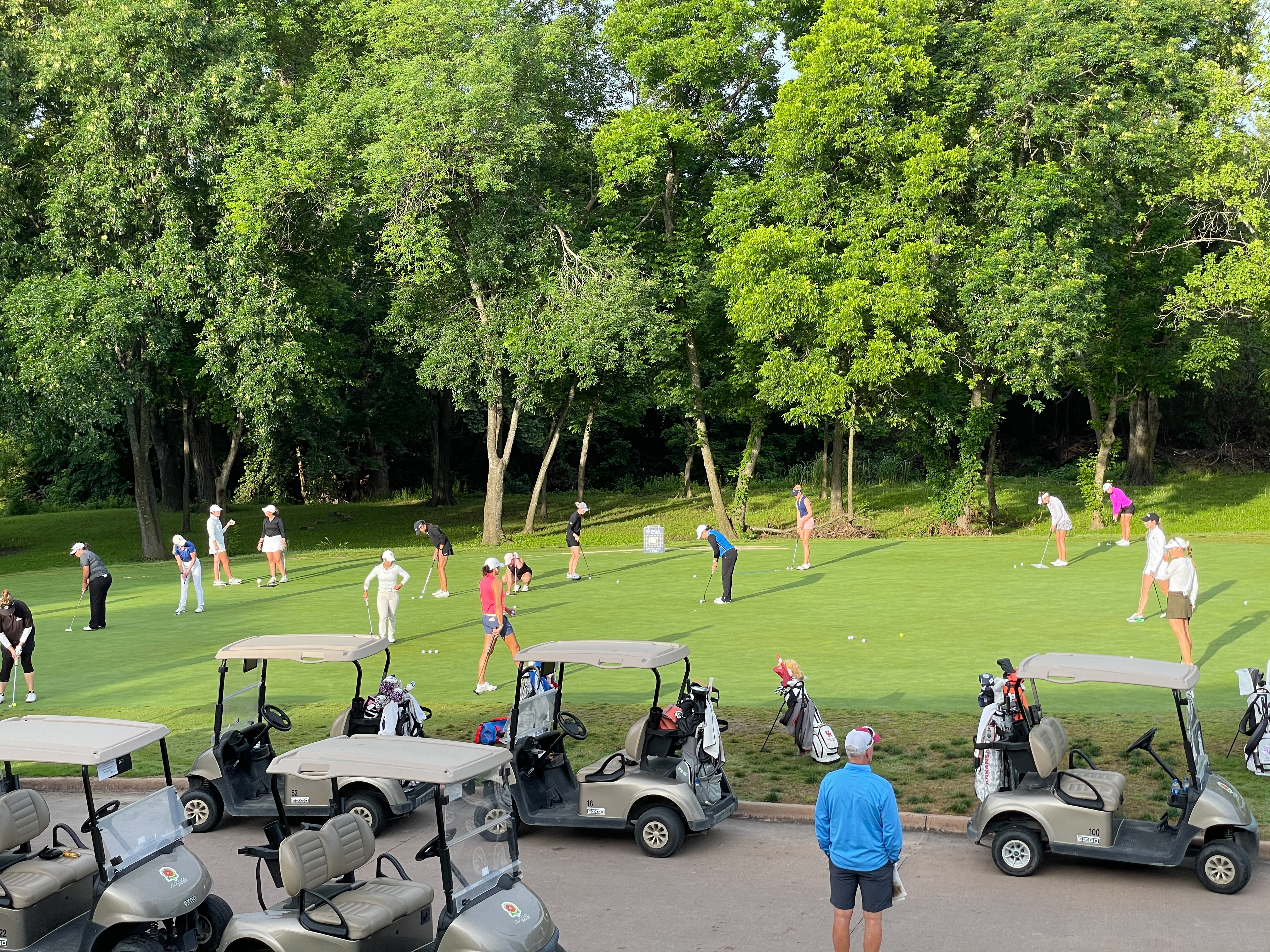 Group of female golfers practicing on the putting green with multiple golf carts parked nearby and trees in the background.