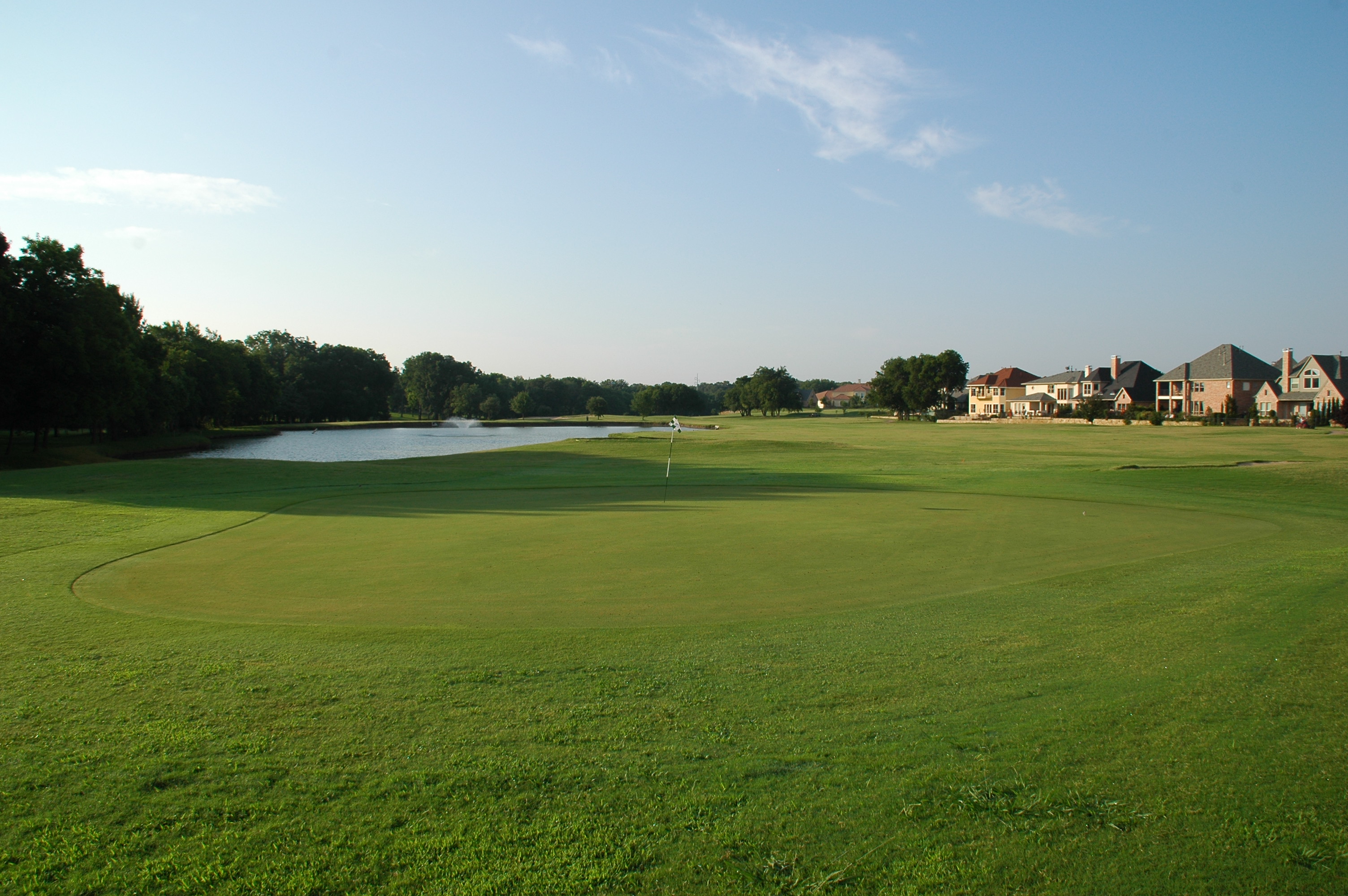 Ground-level view of a putting green with flagstick, surrounded by mature trees and featuring upscale residential homes in the background.
