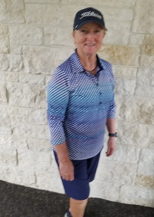 Female golfer in blue patterned shirt and Titleist cap standing against a stone wall background.