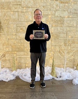 A golfer in dark sweater and glasses standing against the same stone wall holding an award certificate with snow on the ground.