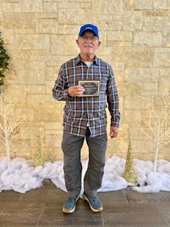 A golfer in plaid shirt and blue cap standing against a stone wall holding an award certificate with snow visible on the ground.