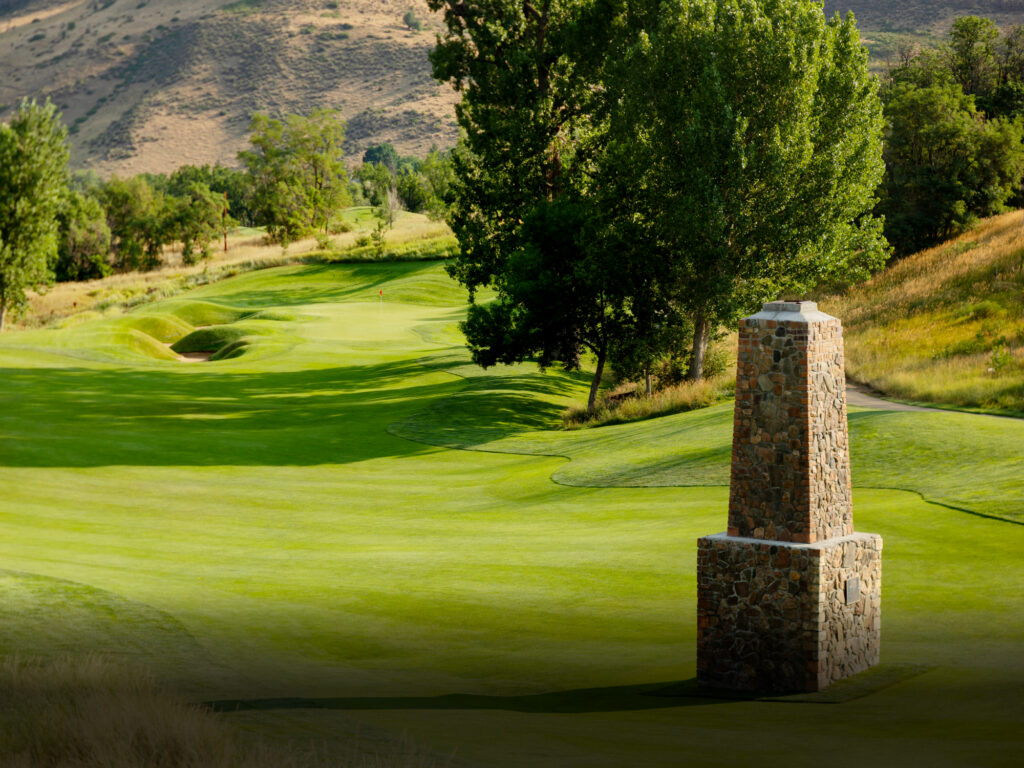 Ground-level view of a golf hole featuring a stone marker monument with rolling hills and desert landscape in the background.