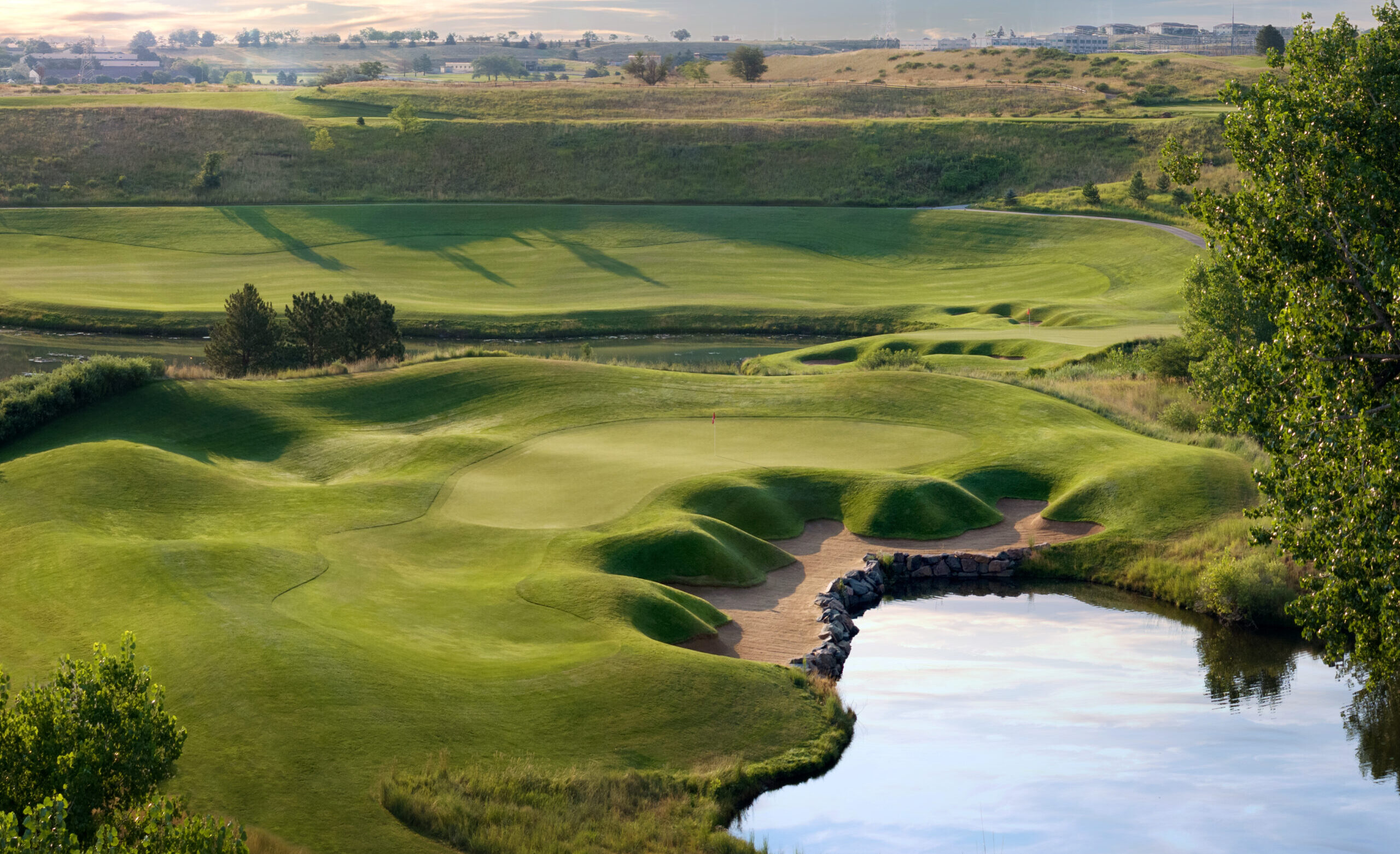 An elevated view of a challenging golf hole with undulating green, strategically placed bunkers, and a water hazard beside the putting surface amid rolling terrain.