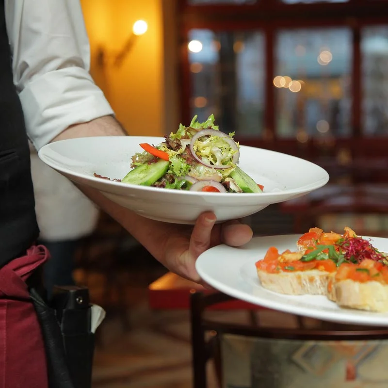 A server presenting elegantly plated dishes including a fresh salad and what appears to be bruschetta or appetizers in an upscale restaurant setting with warm ambient lighting.