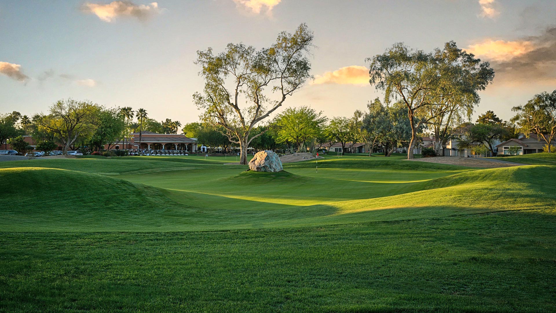 Ground-level view of a putting green during golden hour with a large decorative boulder feature, mature trees, and the clubhouse visible in the background.