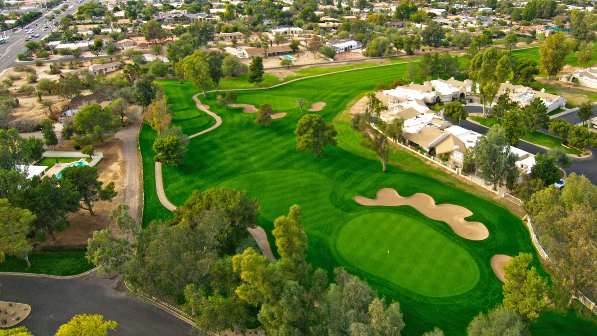 Aerial drone view of a golf hole showing the green surrounded by multiple sand bunkers with residential homes and mature trees in the background.