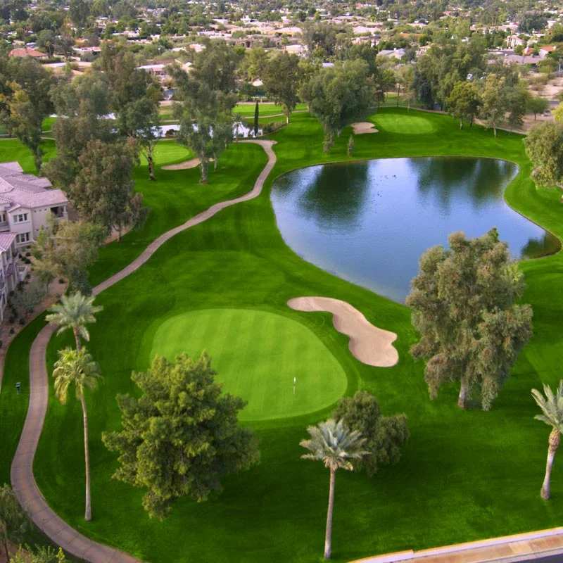 Aerial view showcasing a picturesque golf hole with a large pond water feature, well-manicured fairways and greens, sand bunkers, and surrounding residential neighborhood with mature landscaping.
