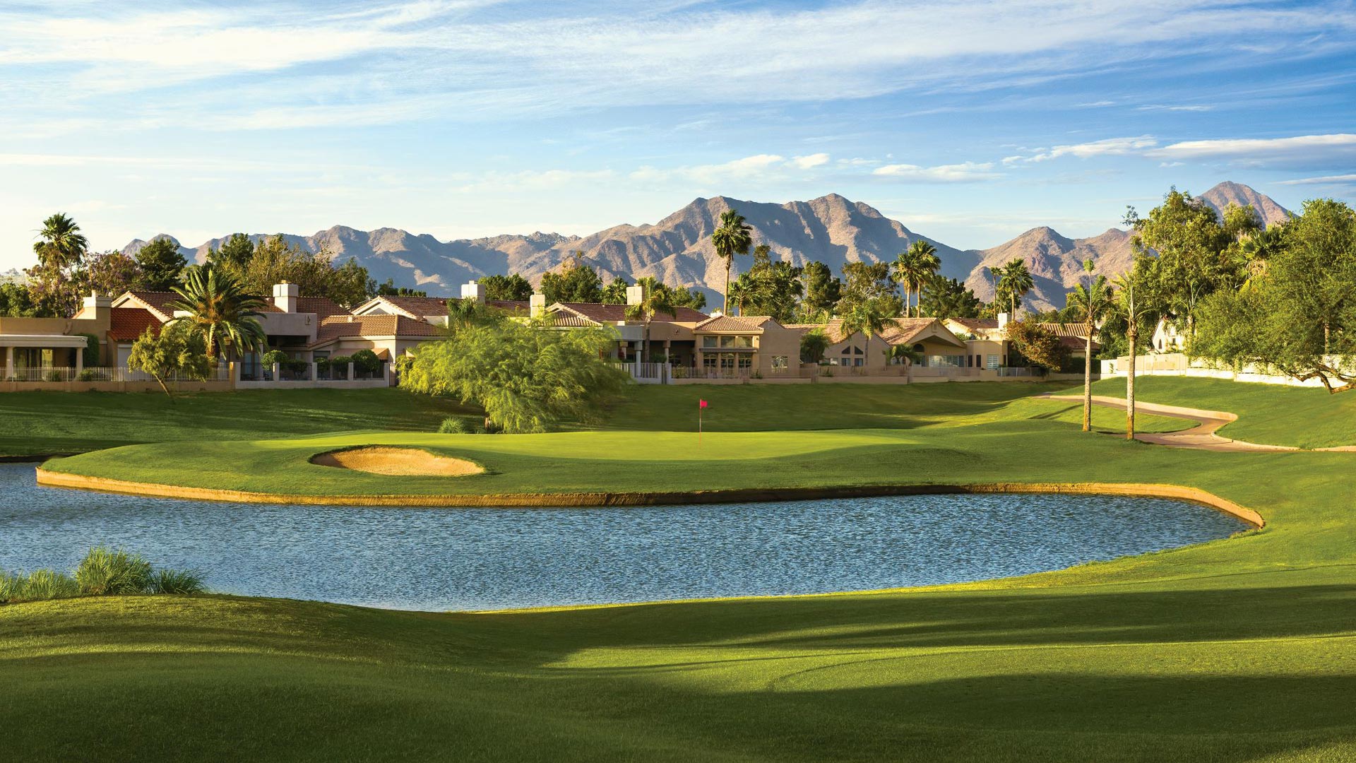 Stunning sunset view of a golf hole featuring a large water hazard that curves around the green with sand bunkers, backed by dramatic desert mountains and upscale residential homes.