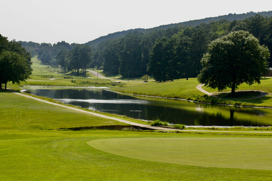 Scenic golf course view featuring a winding water hazard with cart path alongside fairways and rolling hills covered in mature trees.