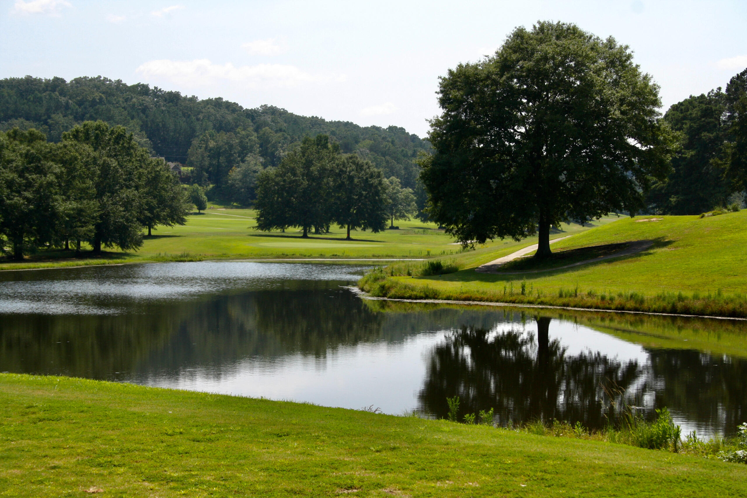A tranquil golf course water feature with perfect reflections of trees and clouds, surrounded by well-maintained fairways and mature landscaping.