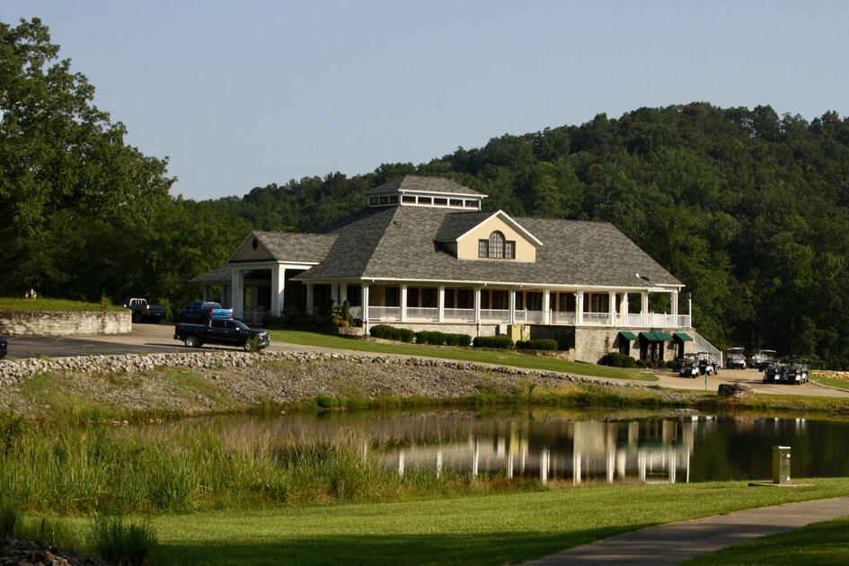Heritage Golf's elegant clubhouse with wraparound porch and stone foundation reflected in the adjacent pond, surrounded by wooded hills with golf carts and vehicles visible.
