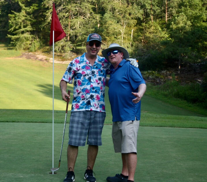 Two golfers posing together on a green with one holding a putter and a red flag visible behind them, surrounded by trees.