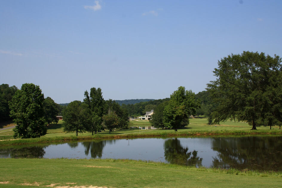 Tranquil golf course pond reflecting surrounding trees with fairways and clubhouse visible in the distance under partly cloudy skies.