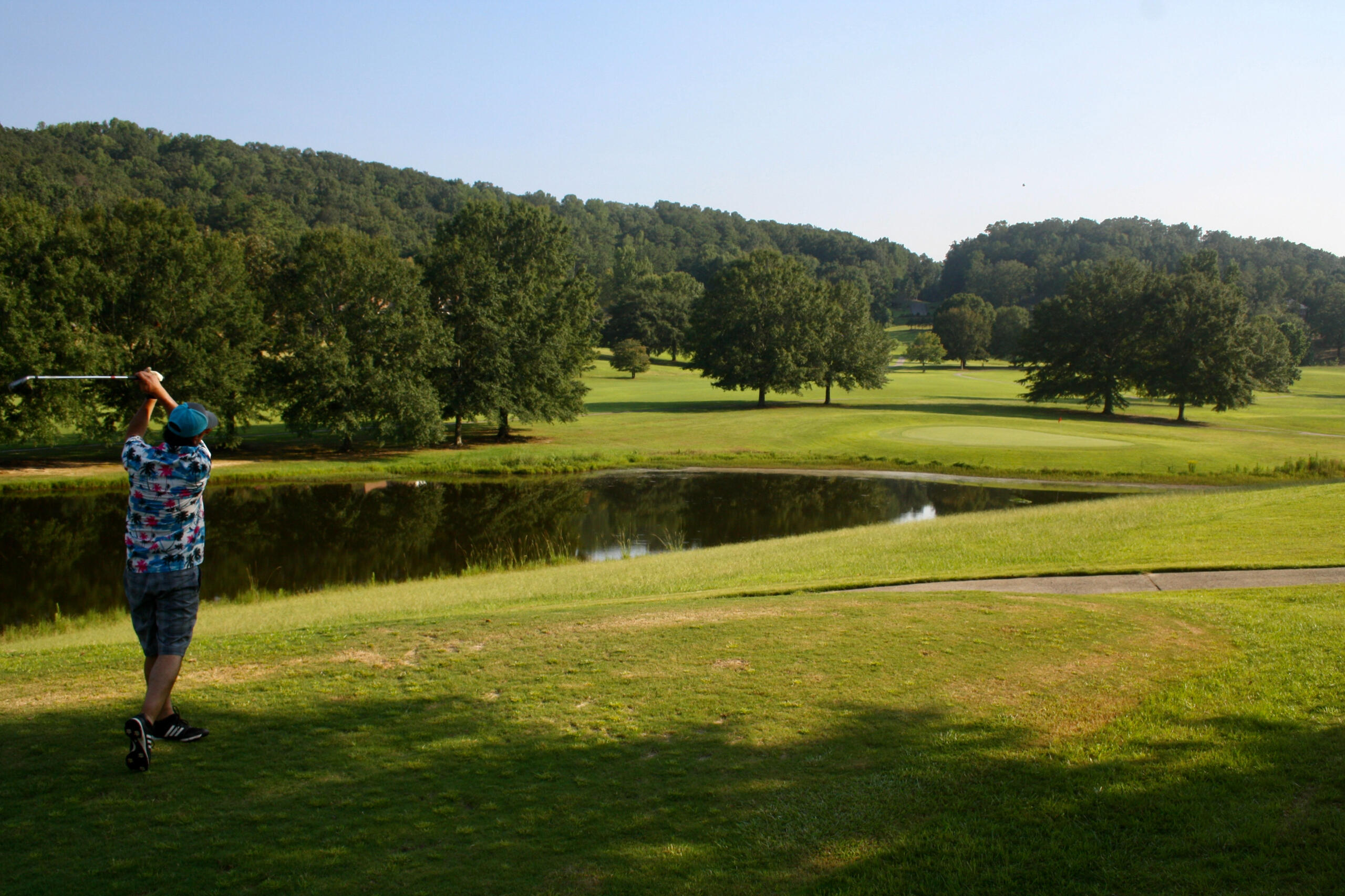 A golfer in a colorful floral shirt preparing to hit a shot near a water hazard with rolling fairways and forested hills in the background.