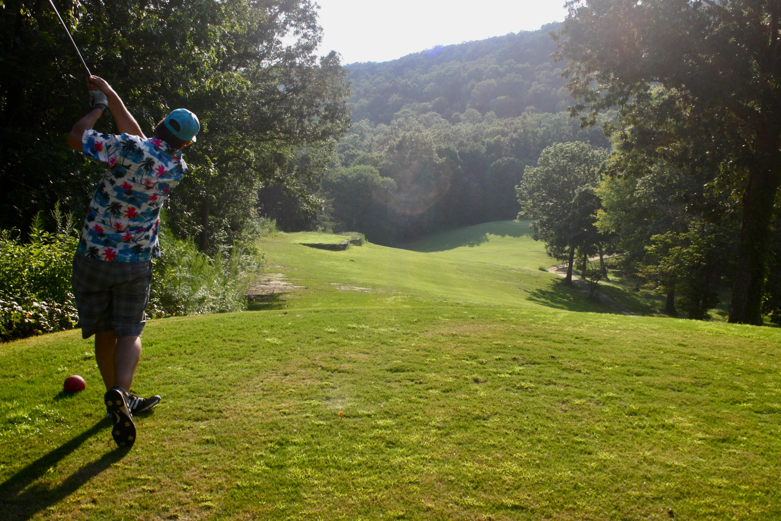 A golfer in colorful attire taking a shot from an elevated tee box with a scenic fairway and wooded valley visible below in warm lighting.