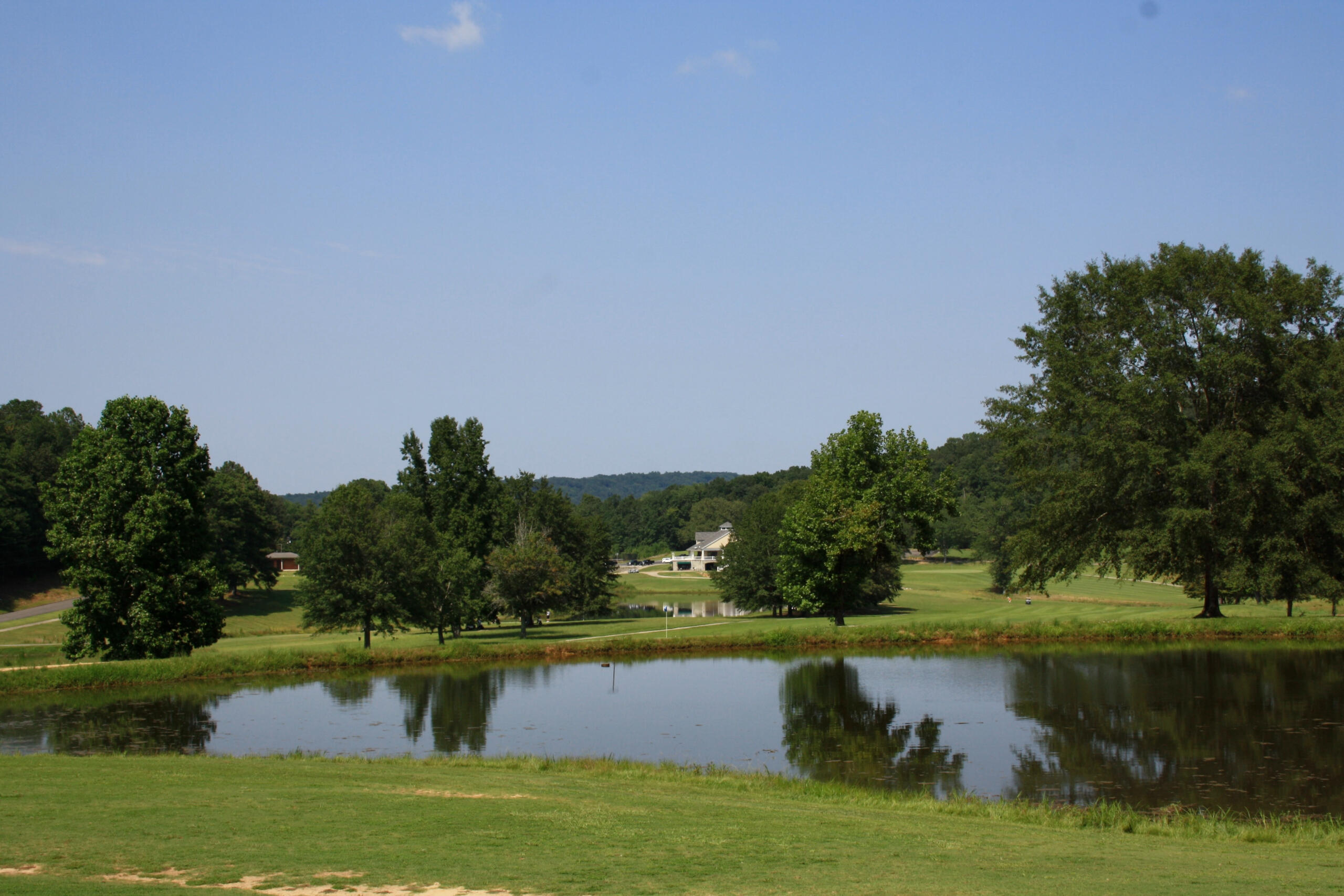 A scenic golf course view featuring a water hazard with reflective surface surrounded by lush green fairways, mature trees, and rolling hills under a partly cloudy blue sky.