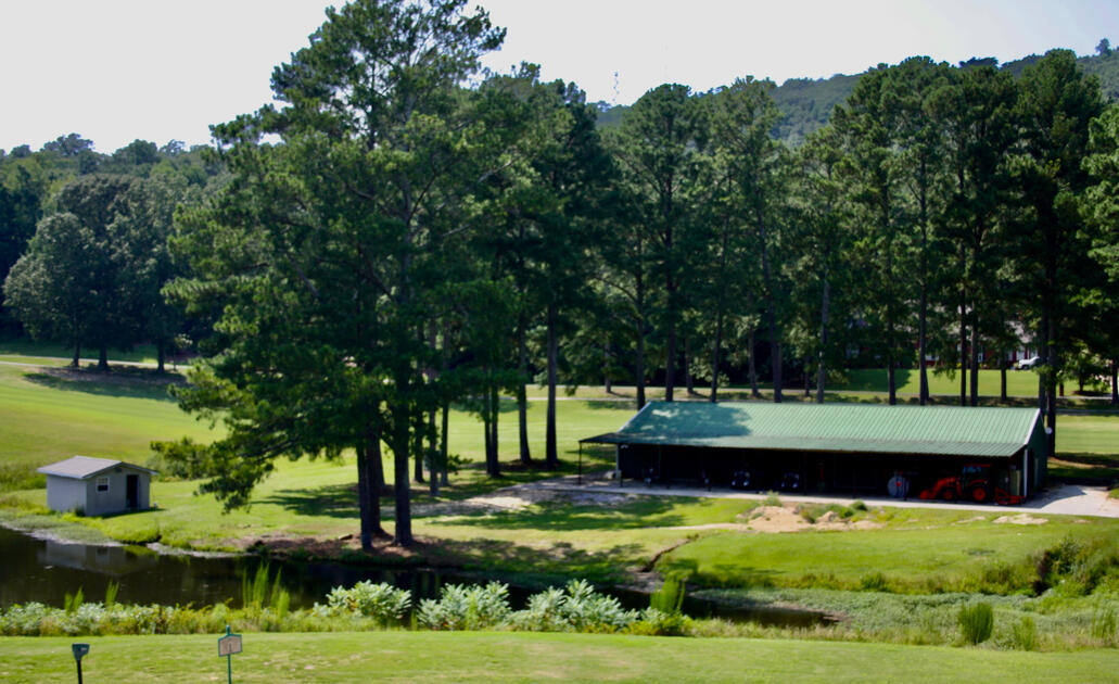 Golf maintenance facility building with green metal roof situated among trees near a water feature and cart storage area.