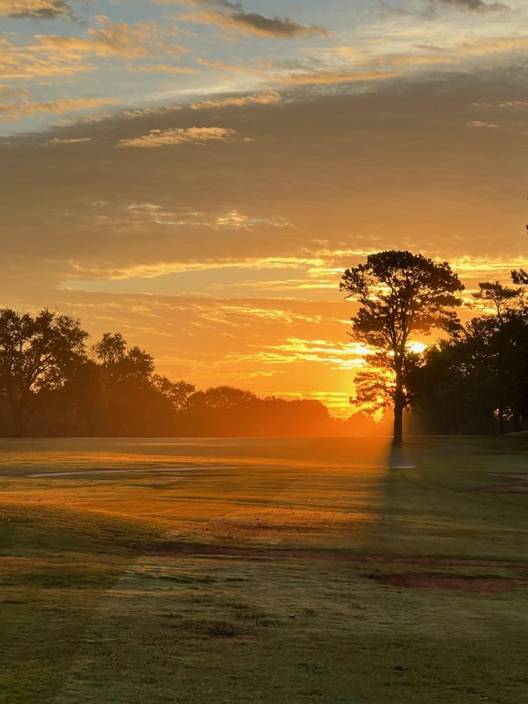 Spectacular golden sunset over the golf course with silhouetted trees and warm orange sky reflecting across the fairway landscape.