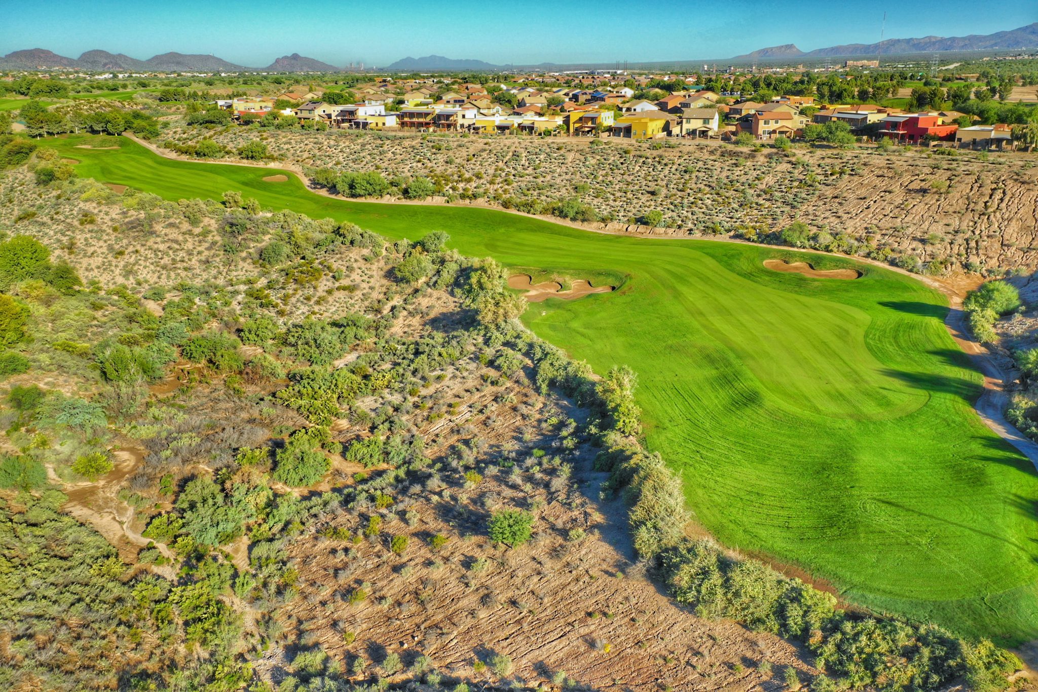 Aerial perspective of a challenging desert golf hole with vibrant green fairway contrasting against natural desert landscape and luxury homes visible in distance.