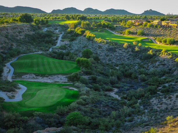 Scenic view of multiple golf holes winding through desert terrain with mountains in the background during golden hour lighting.