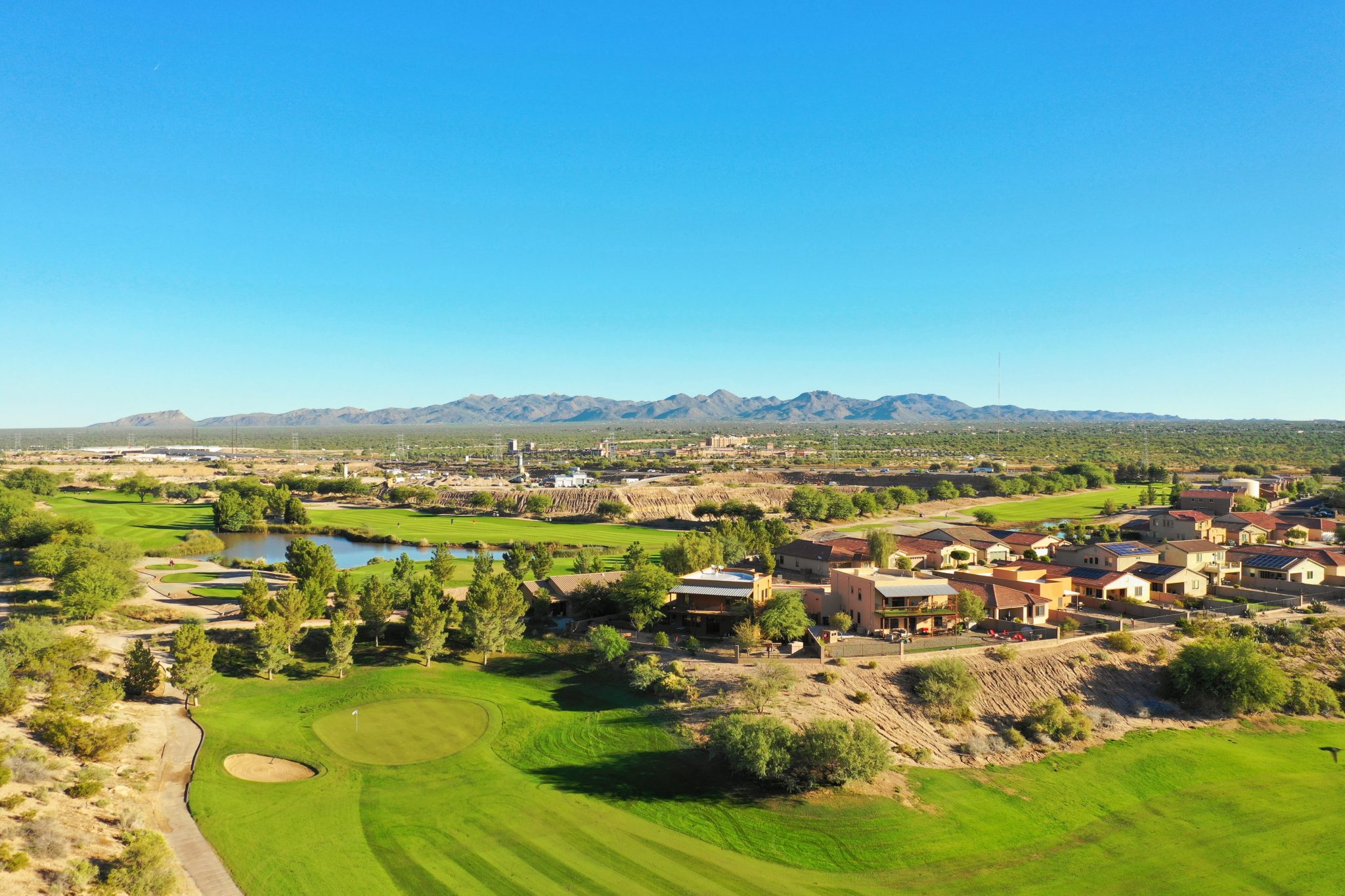 Close-up view of an immaculate putting green with a blue flag pin, surrounded by desert landscape and trees under clear blue skies.