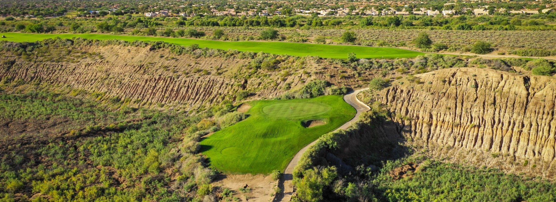 Stunning aerial view of a golf hole carved through dramatic desert terrain with eroded cliff formations and residential development in the background.