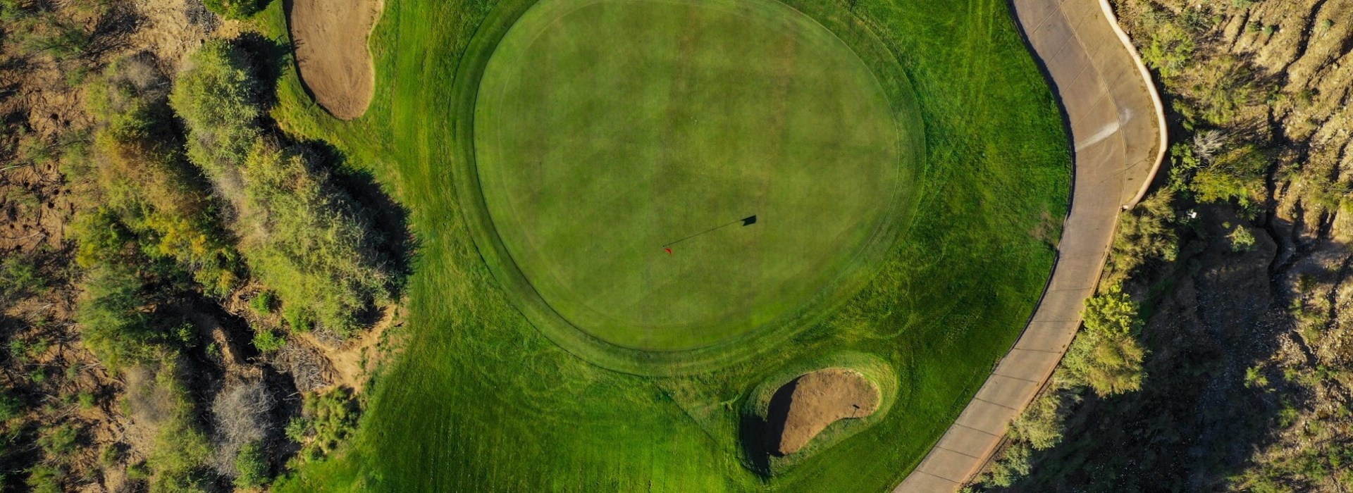 Aerial view of a circular green surrounded by desert landscape with cart path and strategic bunker placement visible.