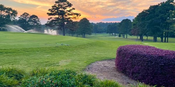 A stunning sunset view over manicured fairways with irrigation sprinklers active, featuring mature trees and well-maintained landscaping including decorative shrubs.