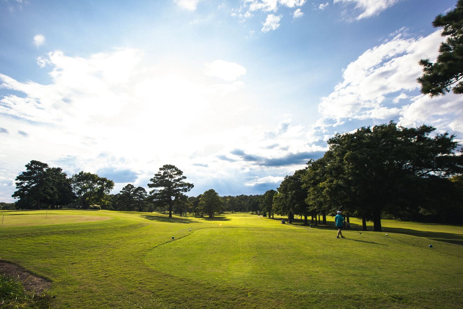 Wide view of the golf course driving range or practice area with a golfer practicing, showing expansive fairways lined with mature trees under a partly cloudy sky.