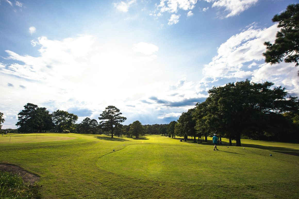 Wide fairway view showcasing the course's open layout with mature trees lining both sides, a golfer visible on the fairway, and dramatic cloudy sky creating an inviting atmosphere for potential members.