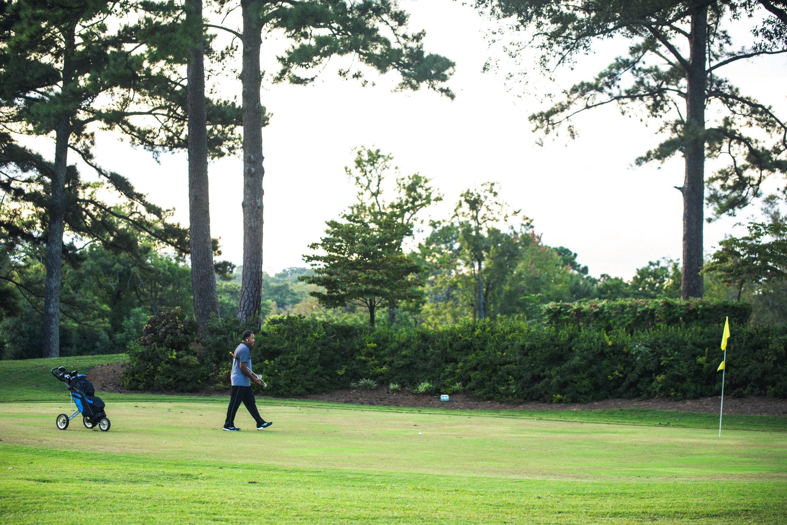 A golfer walking across the fairway pulling a golf cart, with a yellow flag pin visible on the green and mature trees in the background.