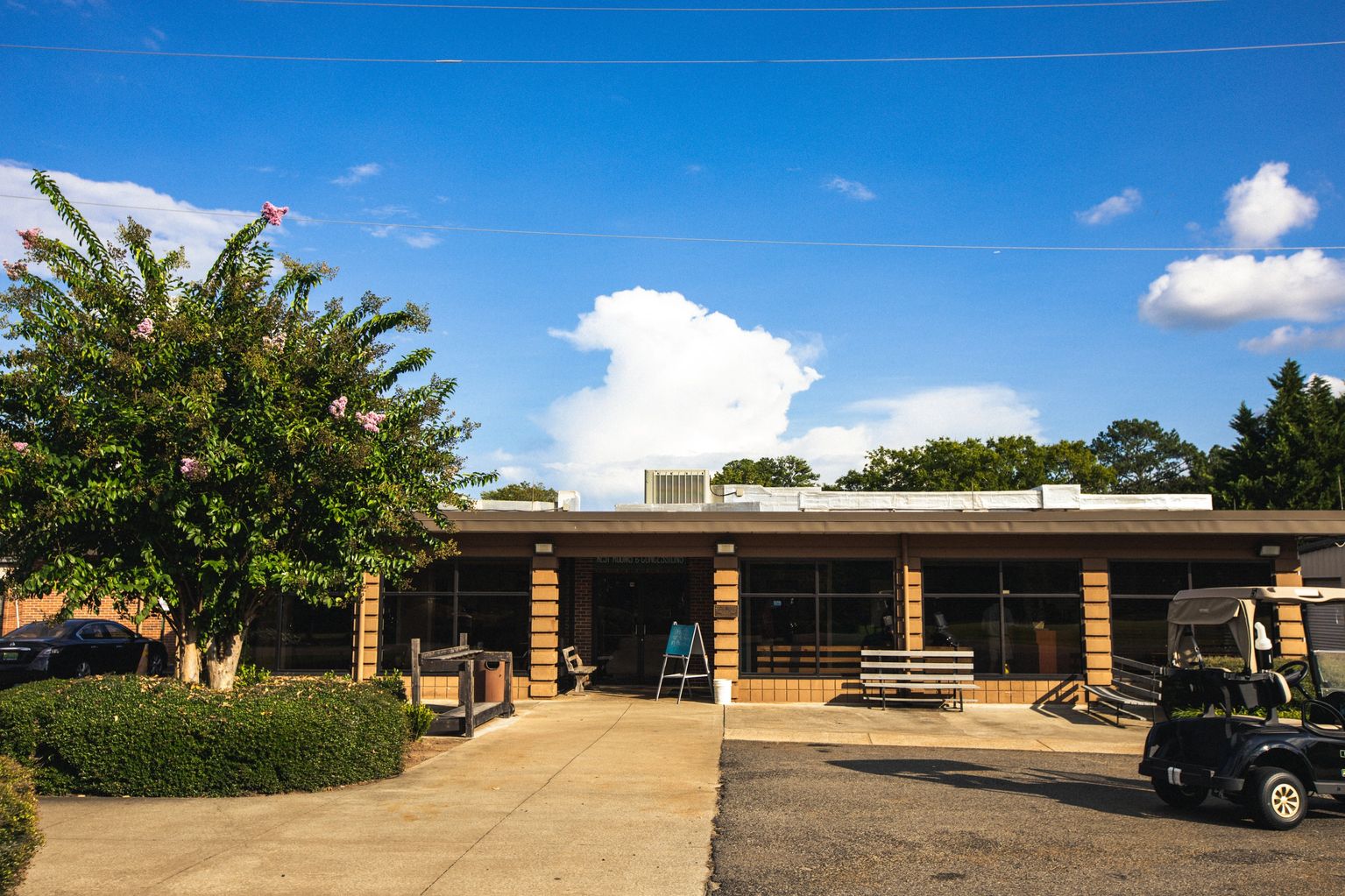 Modern golf clubhouse exterior with brick and wood accents, large windows, and golf carts parked outside under a blue sky with white clouds.