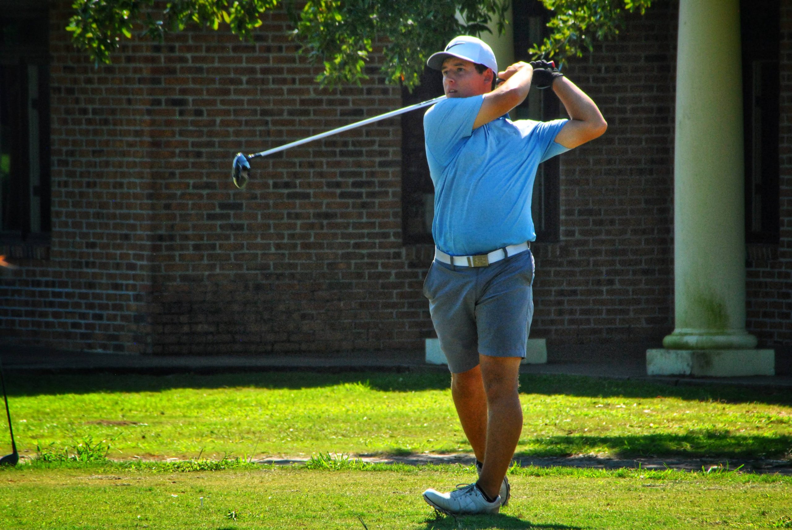 Golfer in light blue attire executing a full swing on the tee with a brick building and green landscaping in the background.