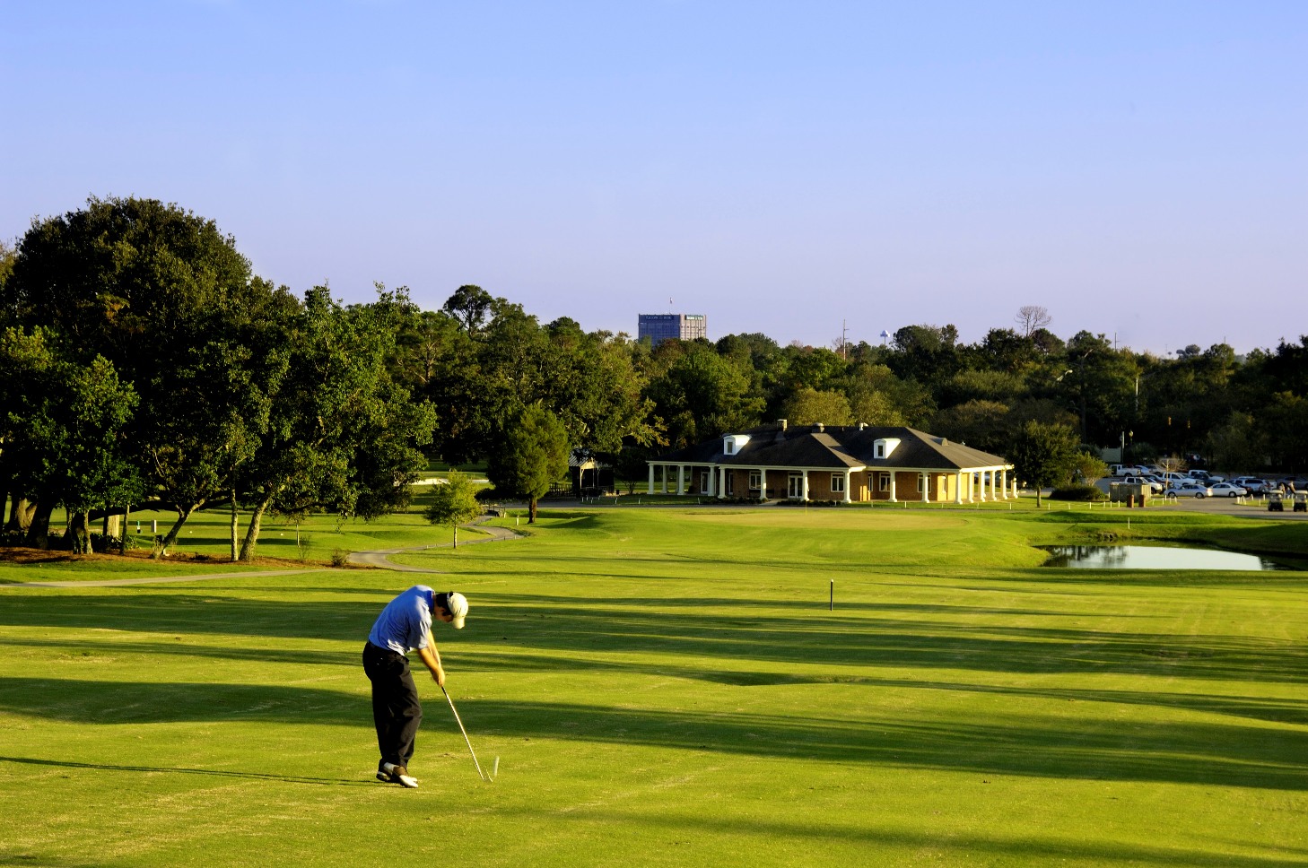 Golfer preparing to putt on a well-maintained green during golden hour with the clubhouse and city skyline visible in the background.