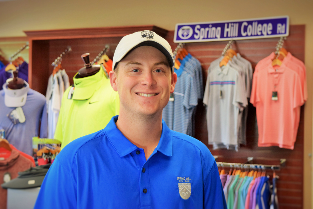 A smiling golf professional in a blue Spring Hill College polo shirt and white cap stands in the pro shop with colorful golf apparel displayed behind him.
