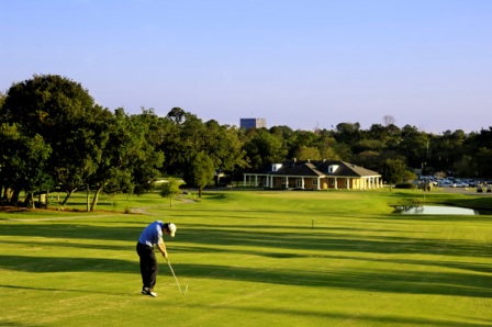 Golfer putting on a green with the clubhouse visible in the background during late afternoon lighting.