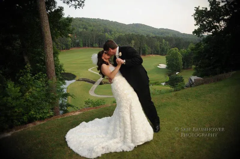 Bride and groom kissing on elevated golf course location with stunning panoramic view of rolling hills, fairways, and forested landscape in background.