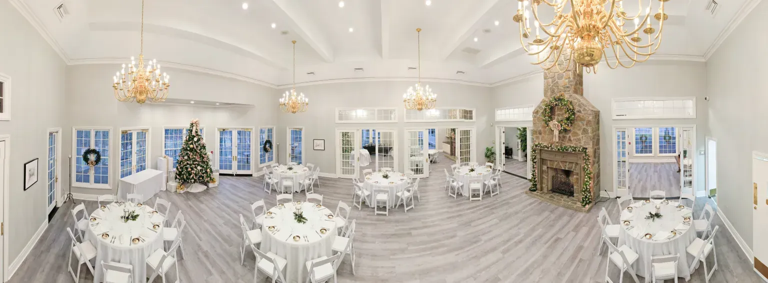 An elegant banquet hall interior with white walls, crystal chandeliers, round tables with white linens, and a stone fireplace decorated for the holidays.