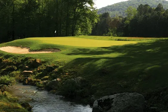 A scenic golf hole with a elevated green surrounded by sand bunkers, a natural stream running through the foreground, and forested hills in the background.