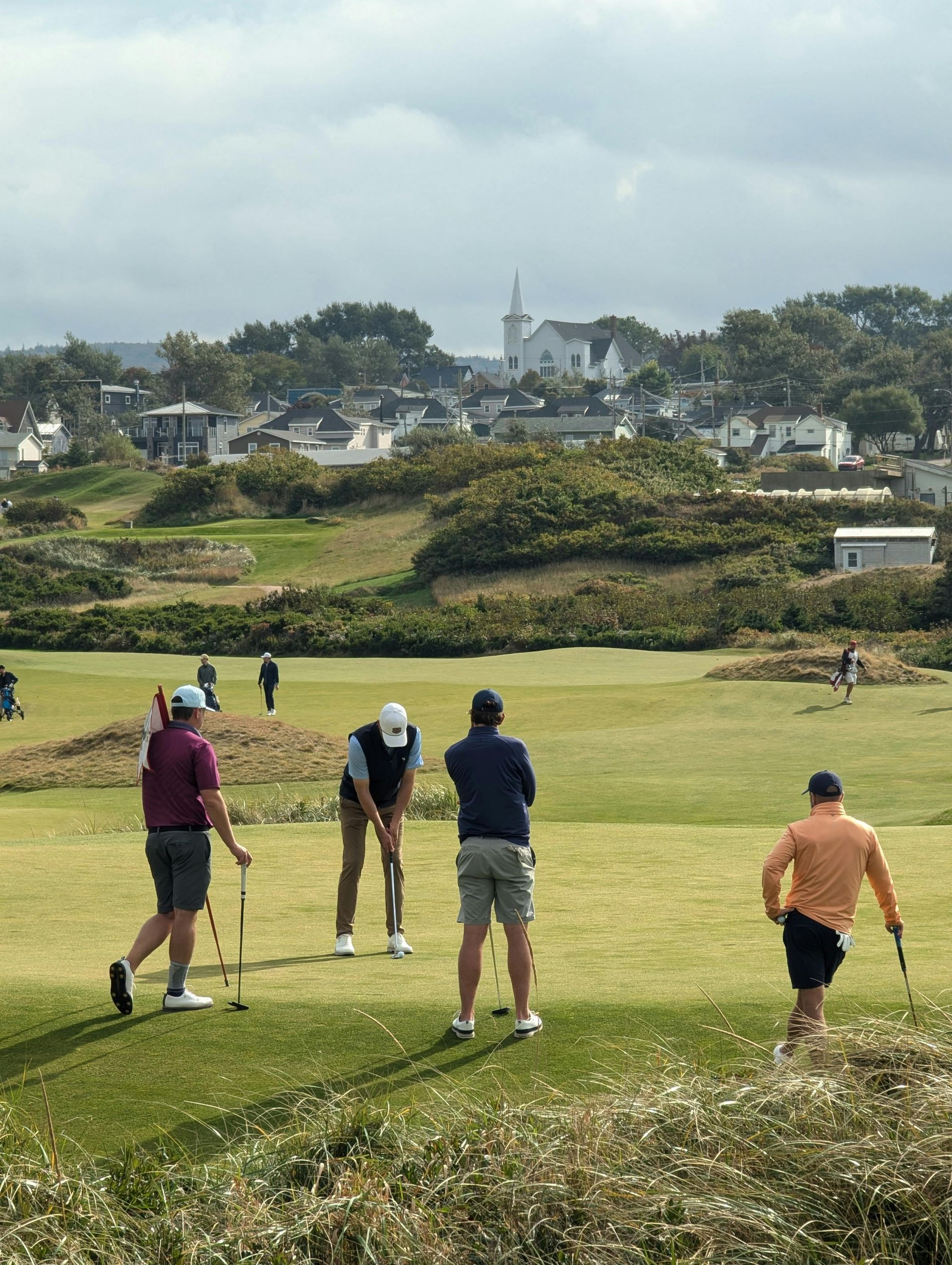 Multiple golfers playing on a links-style course with a white church and residential buildings visible in the background.
