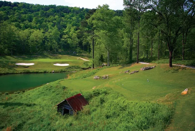Elevated tee shot view showing a challenging hole with water hazard on the left, multiple sand bunkers, and a small rustic structure nestled among mature trees.