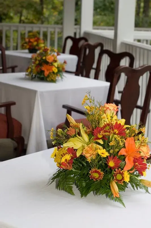 An elegant dining setup on a covered porch with white tablecloths, dark wooden chairs, and autumn floral centerpieces.