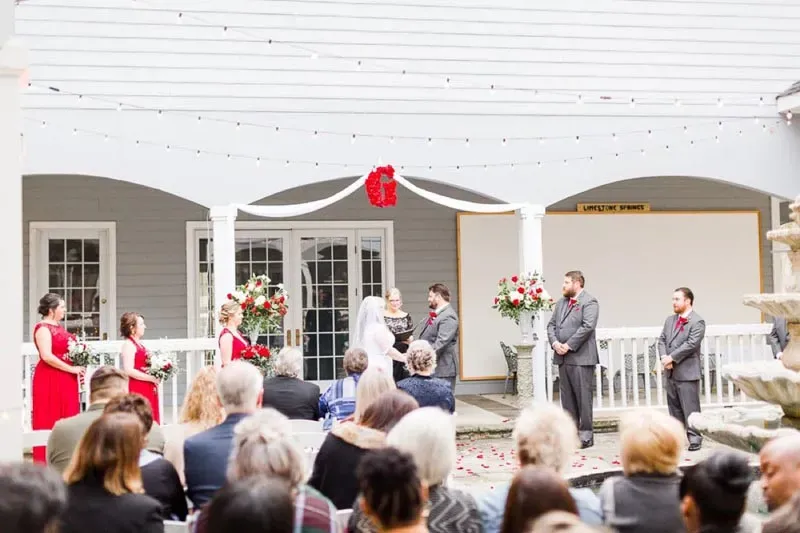 Wedding ceremony taking place on an elegant covered porch with white columns, featuring bride and groom at altar with wedding party and seated guests.